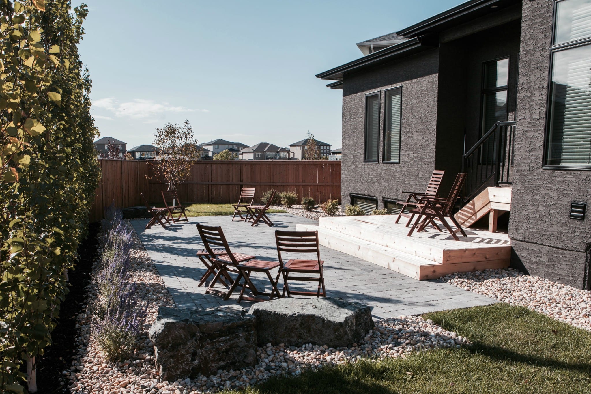 Fully landscaped back yard with a wooden fence, planter beds, rock features, wooden deck and stairs and a brick patio with wooden outdoor furniture.