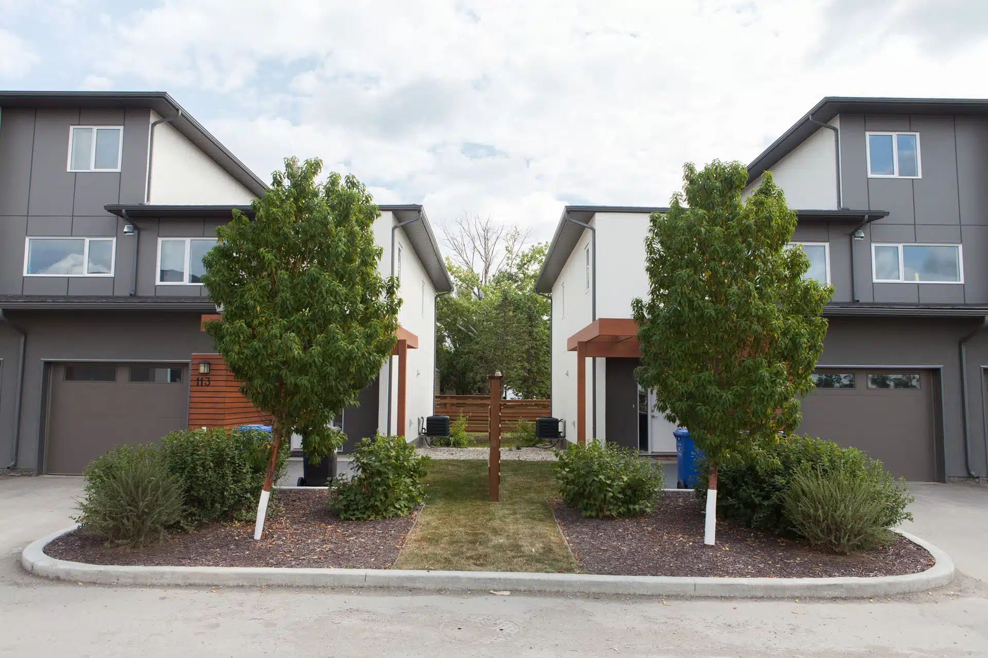 Two modern townhouses with garages, separated by landscaped walkway with trees and shrubs