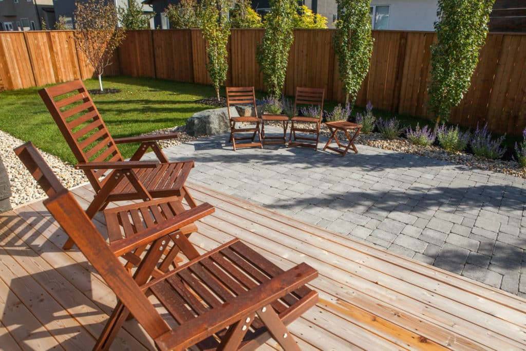 Fully landscaped Winnipeg back yard. Wooden fence and deck, a brick patio, and stone planting bed, with wooden outdoor chairs.
