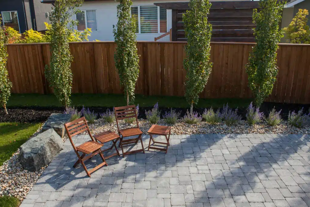 Backyard patio with wooden chairs on pavers, surrounded by landscaping