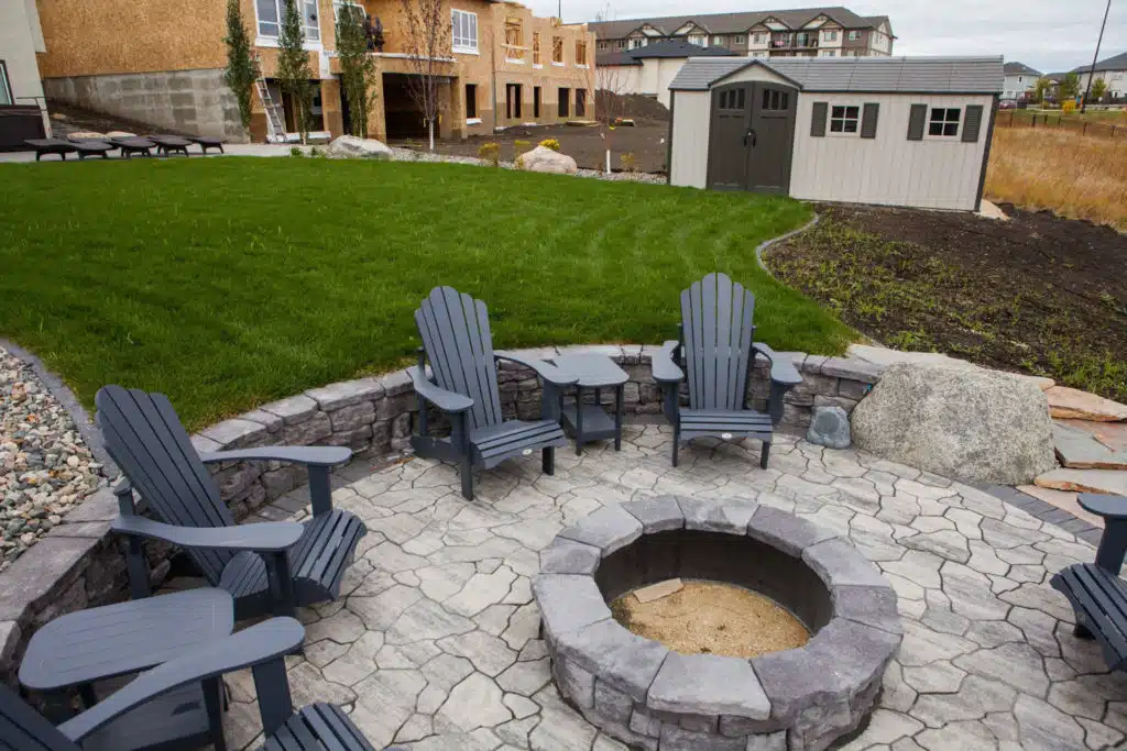 A stone patio with a fire pit encircled by Adirondack chairs and a backyard view