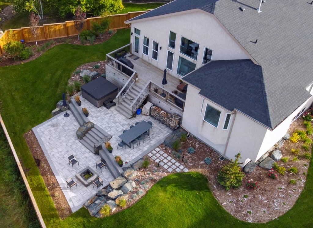 Aerial of landscaped East St. Paul backyard, complete with stone patio & fire pit, elevated deck, rock features and planting beds.