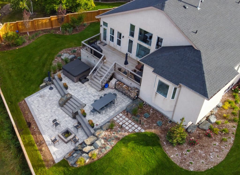 Aerial of landscaped East St. Paul backyard, complete with stone patio & fire pit, elevated deck, rock features and planting beds.