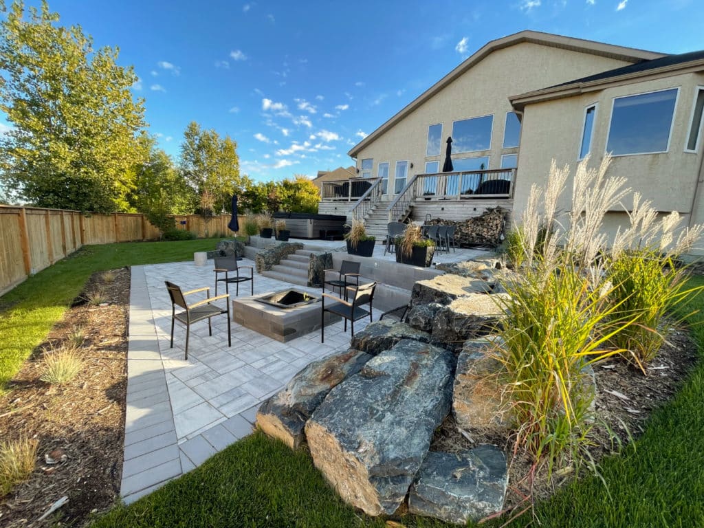 Winnipeg landscaped backyard, with stone patio & fire pit, elevated deck, rock features, planting beds and wooden fence.