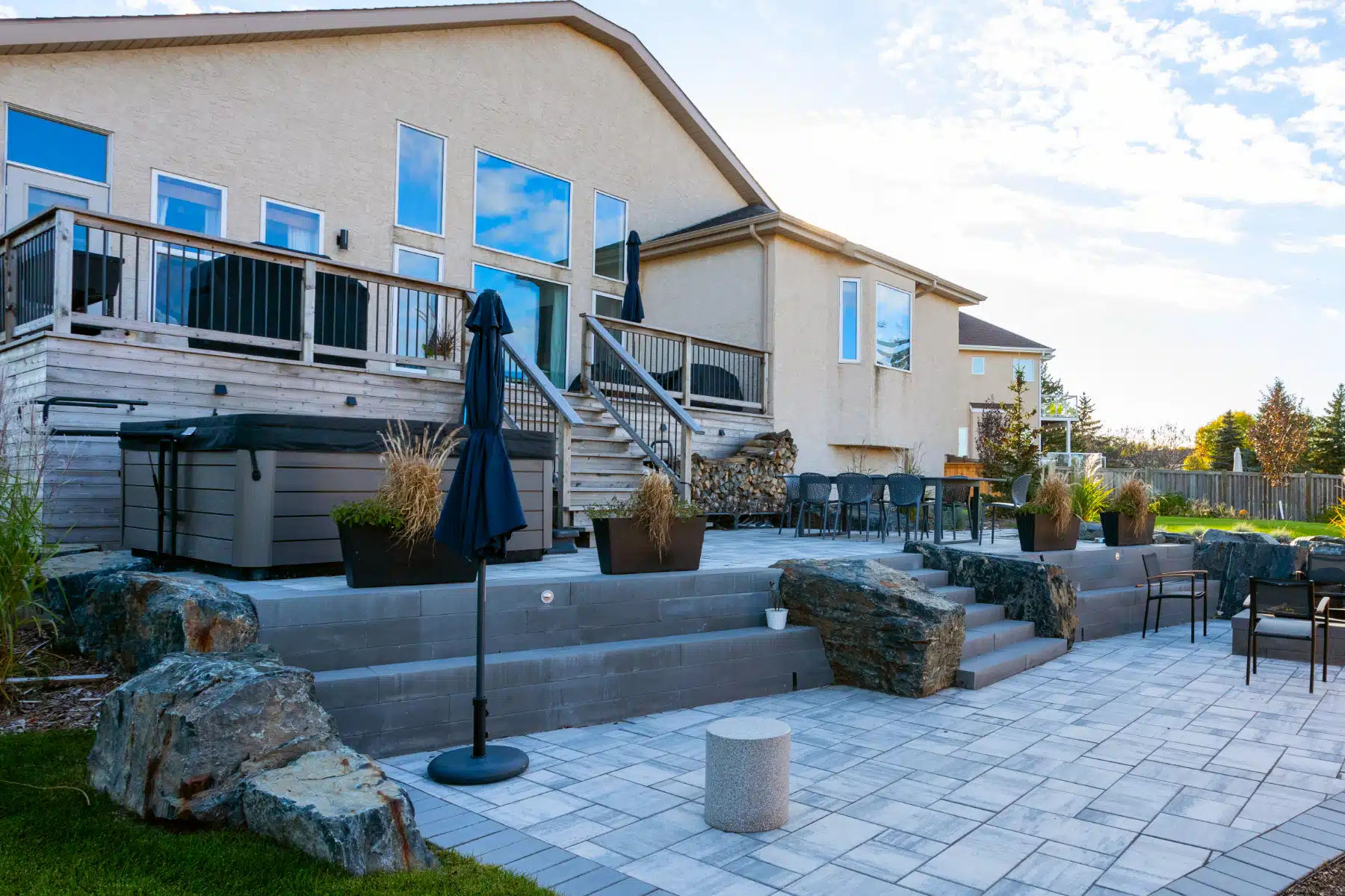Elevated wooden deck, layered stone patio with fire pit and hot tub, and planting beds with rock features, in large East St. Paul backyard.