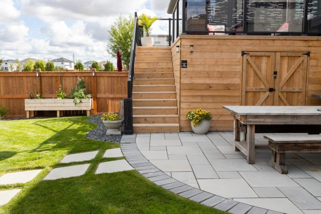 Backyard patio with stone pavers, wooden stairs, rustic dining table, and potted yellow flowers.