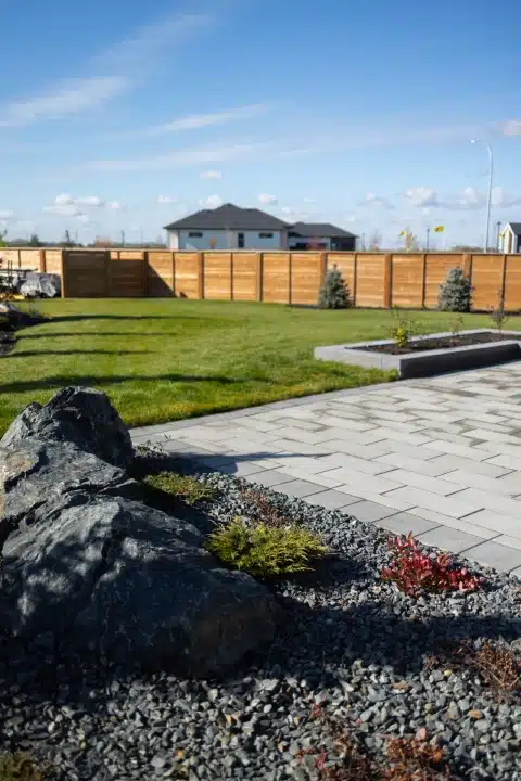 Backyard with a grey paver patio, large boulder surrounded by small shrubs, & wooden privacy fence