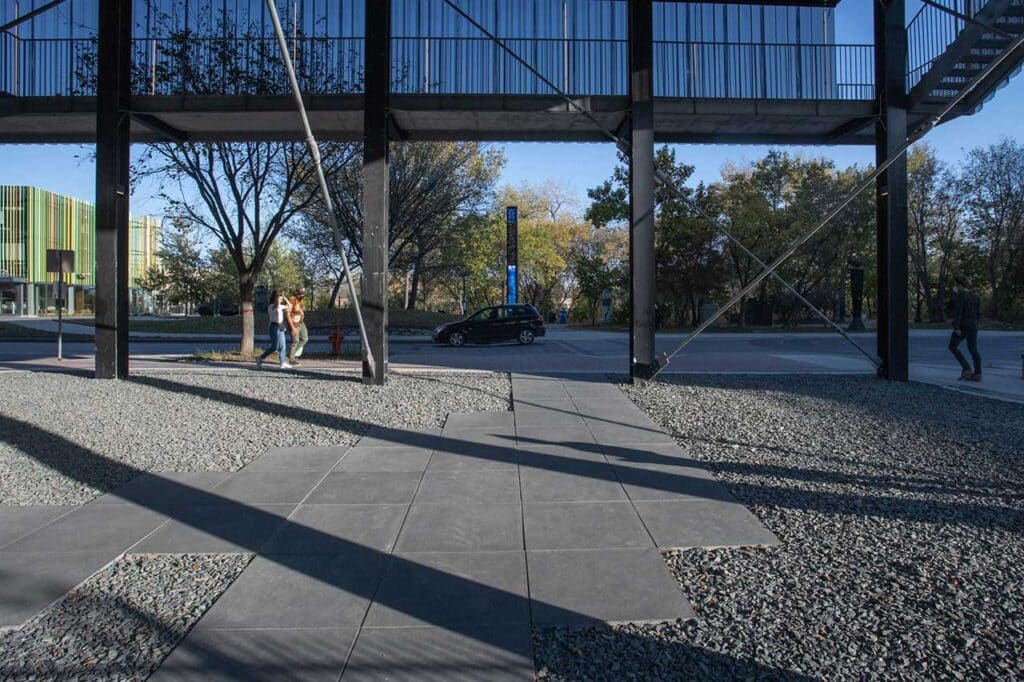 Pedestrians pass under a modern walkway with metal beams, next to a street