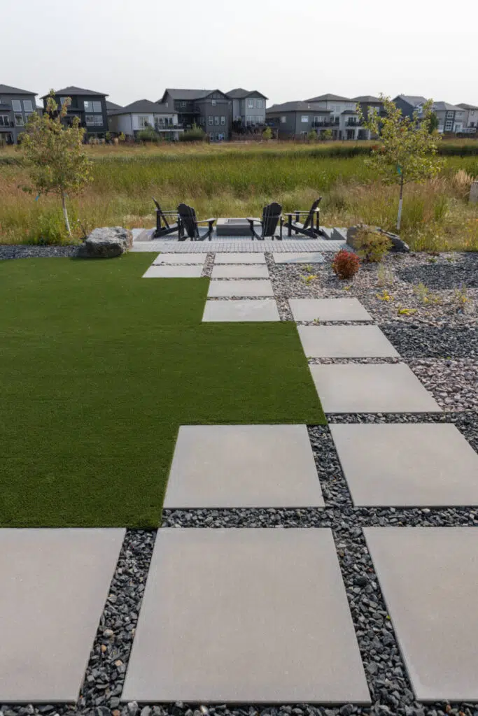 Backyard featuring a path of large square pavers set in dark gravel. vibrant green artificial turf runs alongside the path, leading to a fire pit seating area.