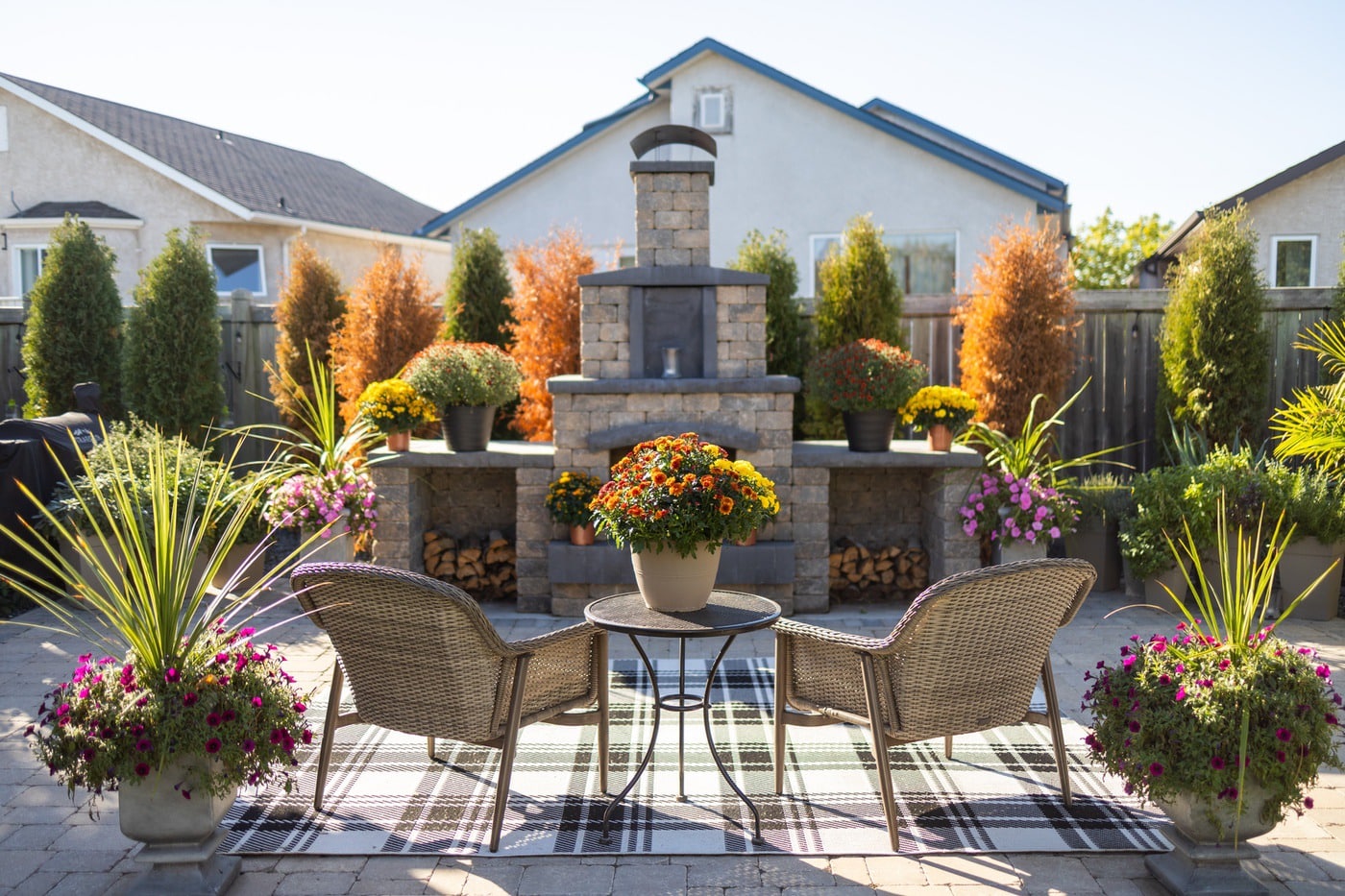 Backyard patio with a stone fireplace, wicker chairs around a small table, and colorful potted flowers including mums and petunias.