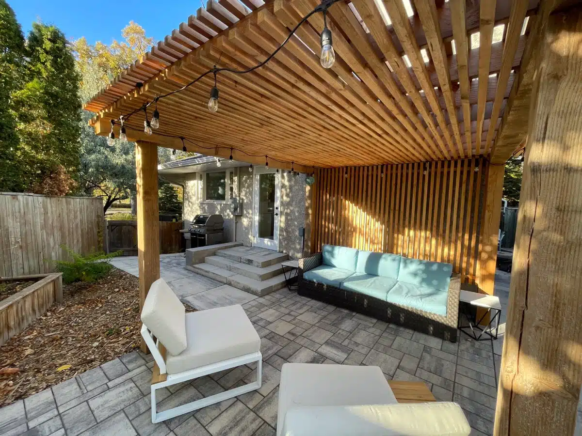 A modern stone patio featuring white armchairs and a blue sofa, all situated under a large wooden pergola with string lights.