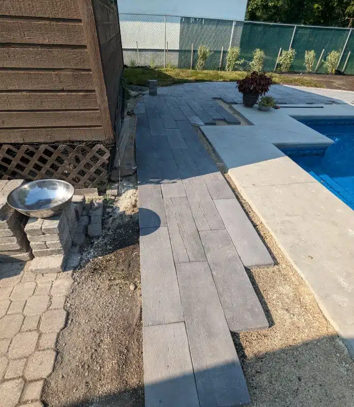 Partially completed backyard poolside walkway with gray plank-style concrete pavers leading alongside a swimming pool.