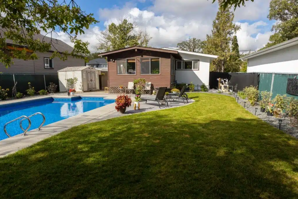 Backyard with a lush green lawn, an in-ground swimming pool, and a patio area in front of a single-story house.