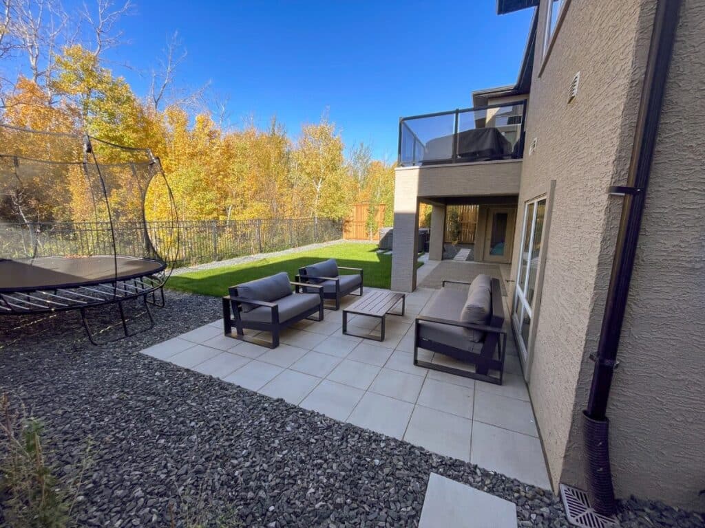 Modern backyard patio with outdoor seating set, tiled flooring, trampoline, and a view of autumn trees along a black metal fence.