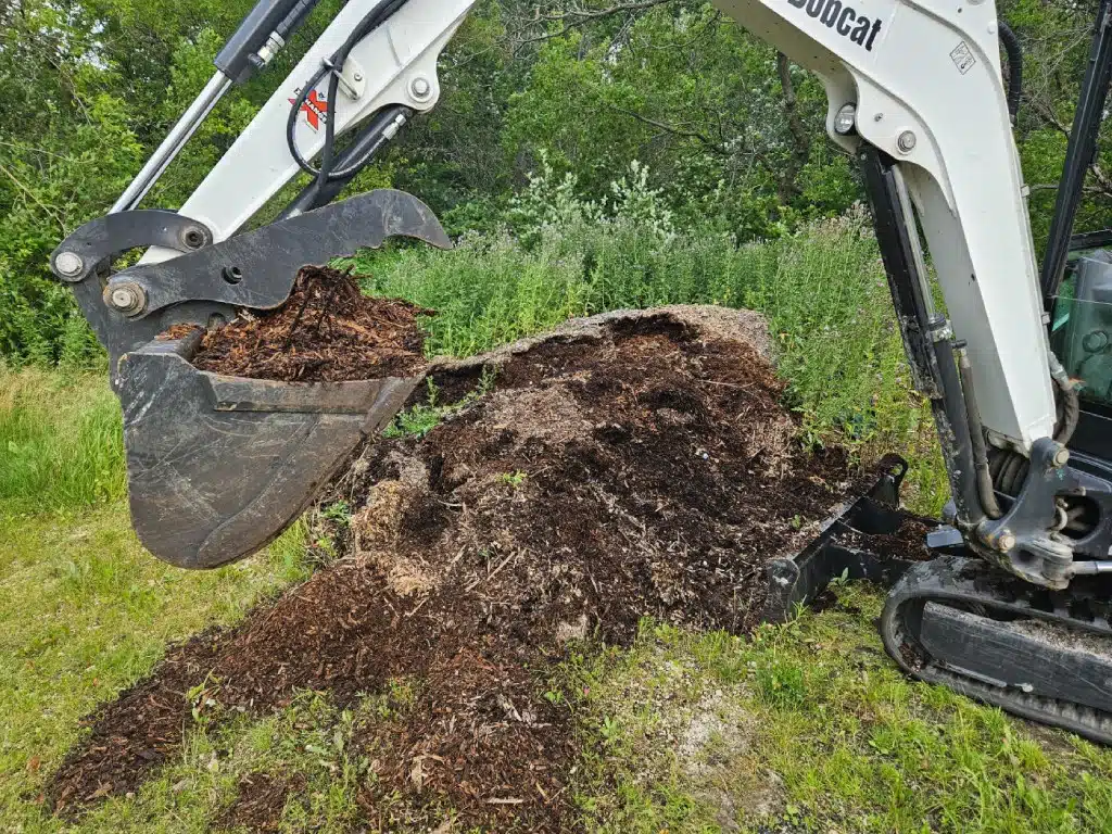 Excavator bucket scooping up a pile of mulch and soil near a grassy area with trees in the background.