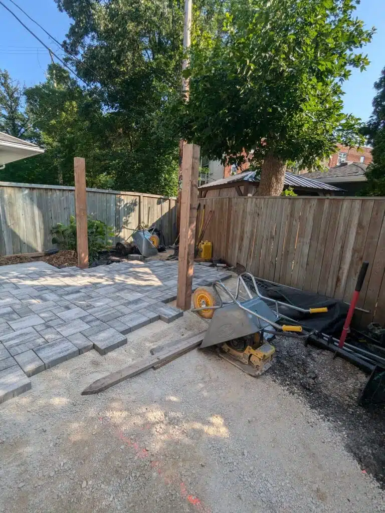 Backyard under construction with paving stones being installed, wooden posts in place, and tools including a wheelbarrow and compactor on site.