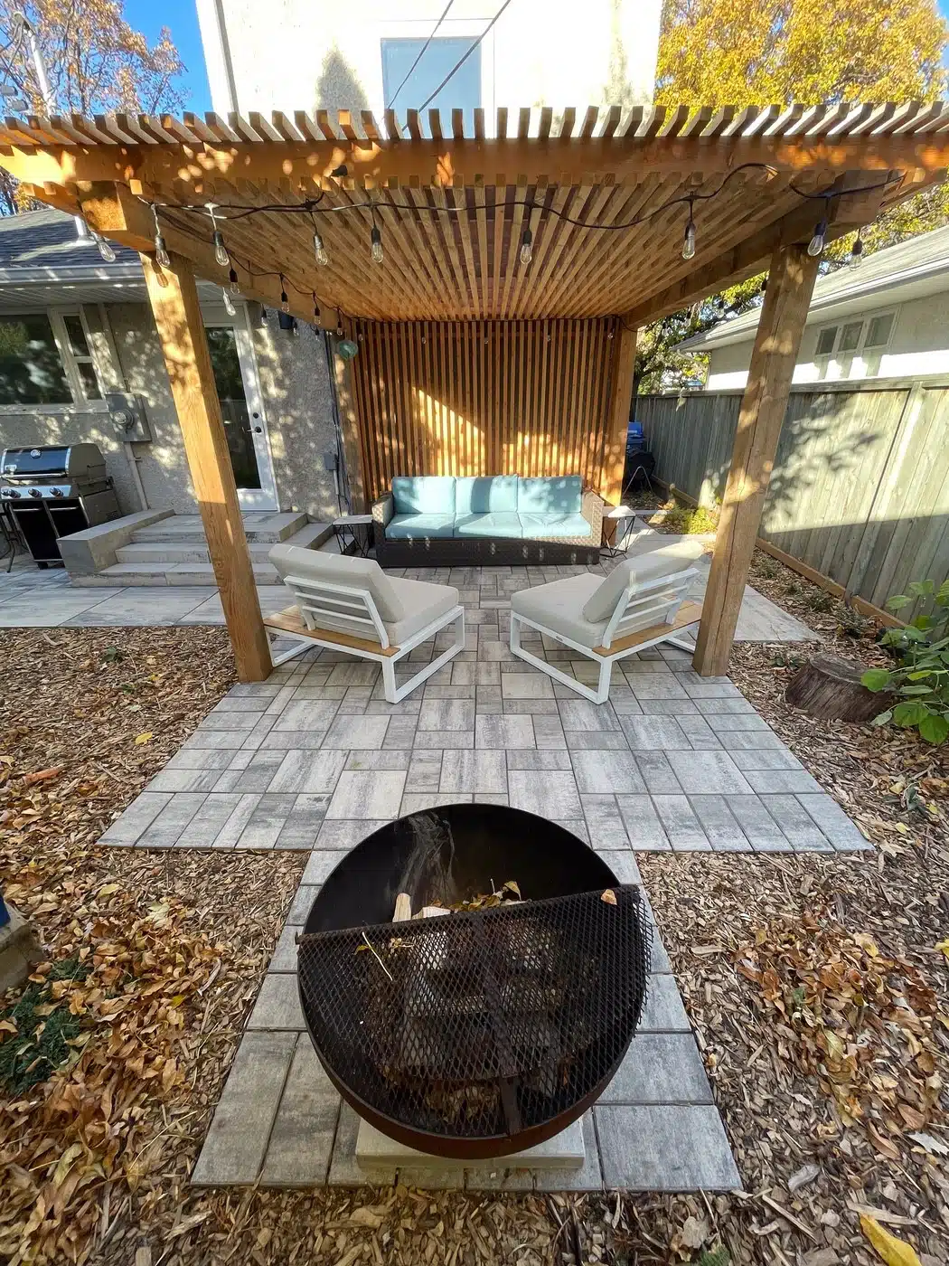 Backyard patio with wooden pergola, cushioned seating, and a round fire pit.