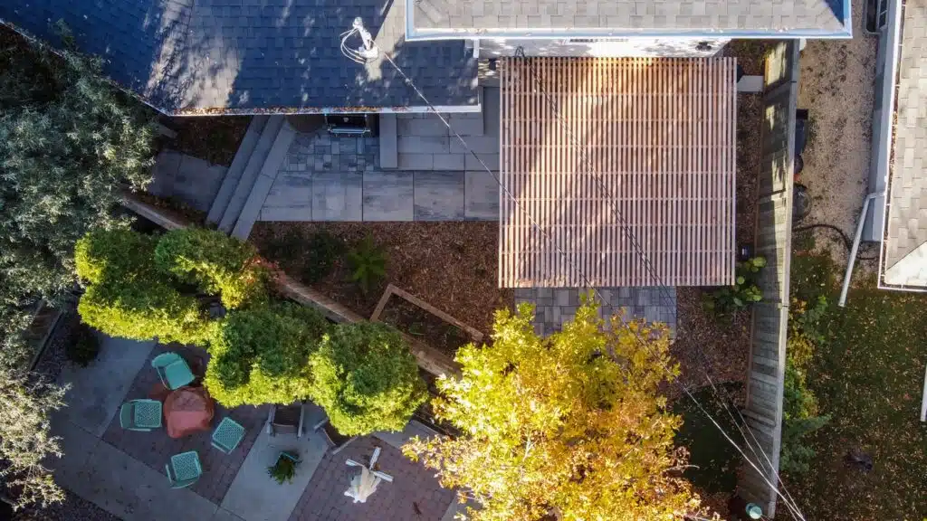 Aerial view of a backyard with a wooden pergola attached to the house, paved patio area with outdoor seating, and trees providing shade.