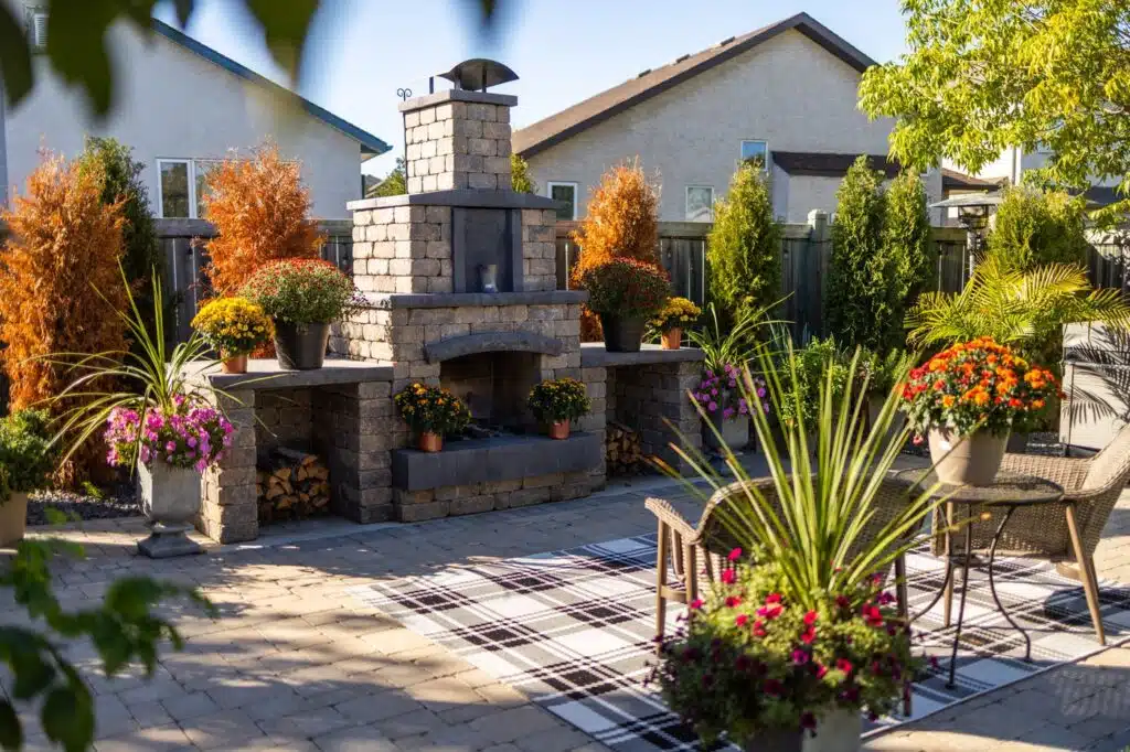 A large brick patio with a large brick fire place, with surrounding flowers, trees and outdoor furniture.