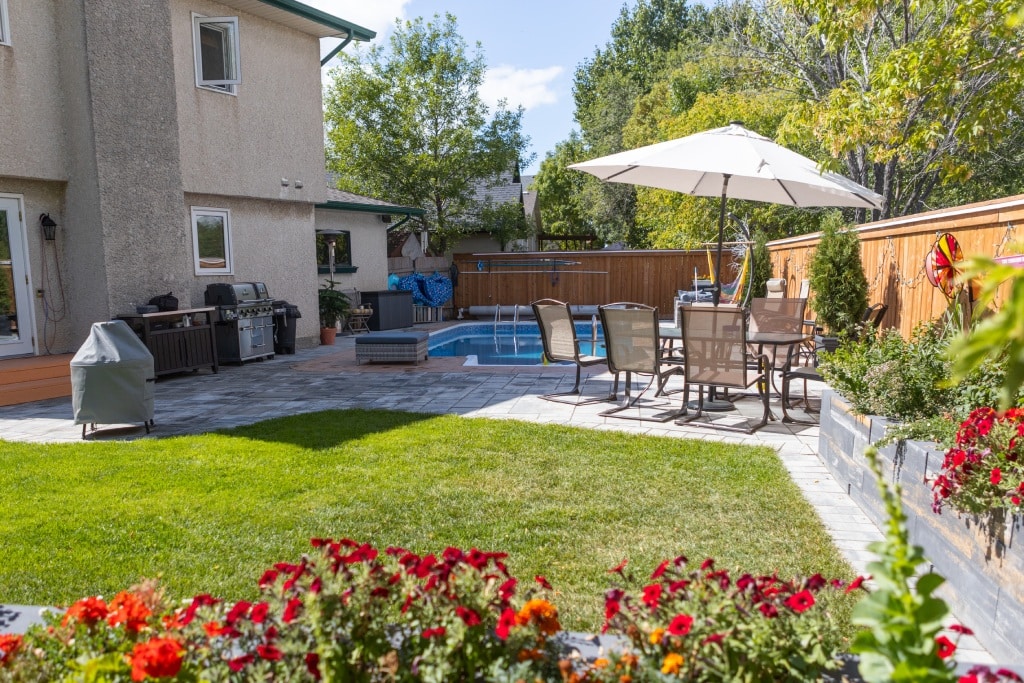 Backyard with a swimming pool, patio dining set under a large umbrella, barbecue area, green lawn, and colorful flowers along the edge.
