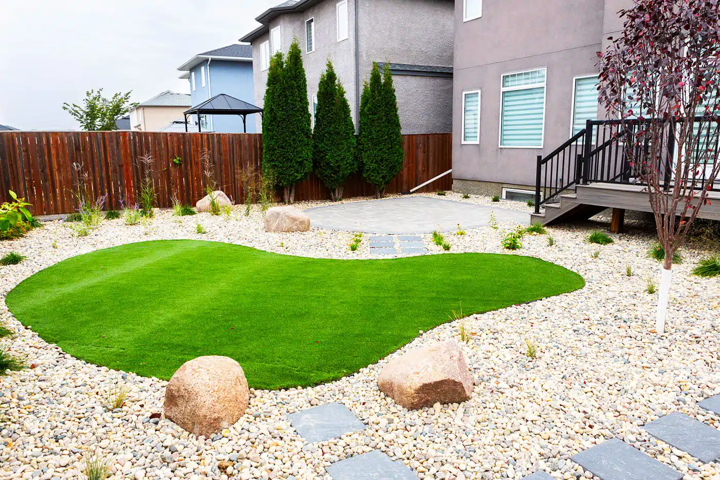 Backyard with a curved patch of artificial grass, stone walkway, and decorative rocks.