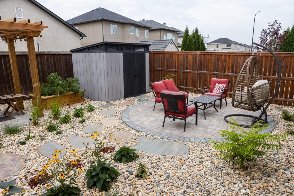 Backyard patio with red cushioned chairs, table, and hanging chair on a circular stone area.